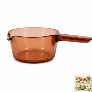 Puede incluir: Un cazo de cristal translúcido de color ámbar con un recipiente redondo, un mango alargado y un pequeño pico vertedor en el borde. El recipiente está fabricado en cristal liso y transparente con un tono marrón cálido en todo el cuerpo.