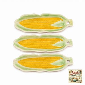May include: Three ceramic corn-shaped serving dishes, each featuring a bright yellow cob design surrounded by light green husks. Each dish has an elongated, textured form with a small circular hole at one end for hanging or display purposes.