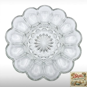 May include: A clear glass deviled egg serving platter featuring a scalloped edge and twelve oval-shaped indentations arranged in a circle. The center of the platter displays a starburst pattern with radiating lines.