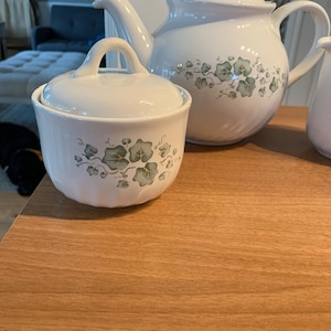 May include: A white ceramic tea set comprising a sugar bowl, teapot, and creamer. The set is decorated with a subtle green and grey ivy leaf design. The teapot has a curved spout and handle, and the sugar bowl has a lidded loop handle.