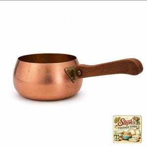 May include: A round copper saucepan with a smooth, metallic finish and a single, curved wooden handle attached by a brass-colored metal bracket. The pan features a wide, open top and a rounded base, showcasing a warm, reddish-brown color palette.