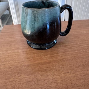 Puede incluir: Una taza de cerámica con asa y base oscuras. La taza tiene un esmalte de dos tonos, que va del verde-azulado al marrón oscuro. La taza está sobre una superficie de madera.