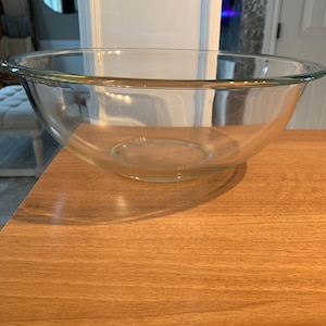 May include: A clear glass mixing bowl. The bowl has a rounded shape and is sitting on a wooden surface. The bowl is empty and clean.