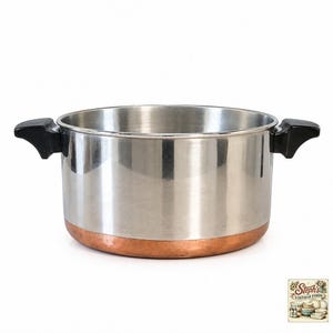 Puede incluir: Una olla de acero inoxidable con una base de color cobre y dos asas laterales negras. La olla presenta un acabado metálico pulido en las paredes exteriores e interiores. Un pequeño logotipo en la esquina dice Steph's VINTAGE FINDS.