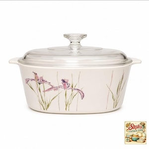 May include: A white ceramic casserole dish with a clear glass lid, featuring a painted floral pattern of purple iris flowers and green stems on the exterior. The dish has two side handles and a round knob on the lid. Includes a small logo graphic.