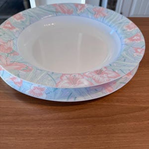 Puede incluir: Dos cuencos de cerámica blanca apilados sobre una superficie de madera. Los bordes de los cuencos están decorados con un estampado floral en rosa, azul claro y verde. Los cuencos tienen un diseño ancho y poco profundo.