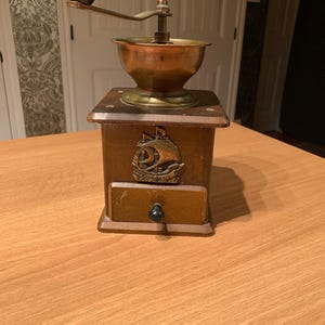 May include: A vintage coffee grinder with a wooden base, brass bowl, and a black handle. The grinder features a drawer and a decorative emblem of a ship. The grinder is sitting on a wooden table.