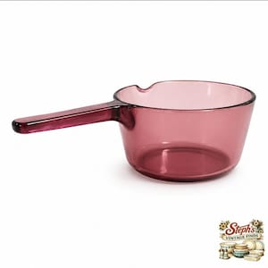 May include: A translucent plum-coloured glass saucepan featuring a round basin and a long, integrated handle. The rim of the vessel includes a small pouring spout indentation on one side. The glass is smooth and uniform in its deep purple hue.
