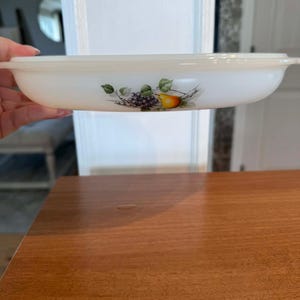 May include: A white oval serving dish with a lid. The dish features a fruit design with grapes, a pear, and green leaves. The dish is being held in front of a white door.