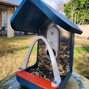 May include: A blue bird feeder with a white plastic perch and a Nest Cam mounted inside. The feeder is filled with birdseed and has a roof to protect the camera from the elements.