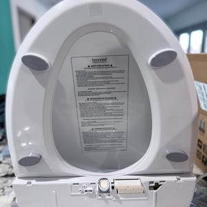 May include: A white Brondell bidet toilet seat with a safety precautions label centered on the underside. The seat features four grey rubber bumpers and a control housing at the base with a circular dial and a rectangular panel with printed text.