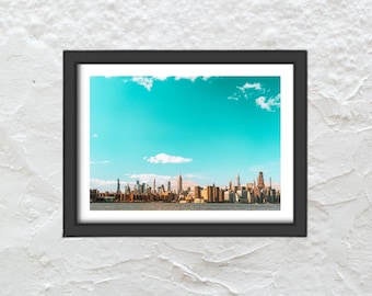 Pode incluir: Uma impressão emoldurada com uma paisagem urbana sob um céu turquesa brilhante com nuvens brancas. A moldura preta envolve a imagem do horizonte da cidade, que é definida contra um céu azul vibrante.