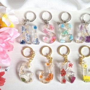 May include: Resin letter keyrings with dried flowers and gold leaf inclusions. Each letter is attached to a silver or gold keyring. The letters include M, S, E, A, G, J, K, and L. The background includes pink, yellow, and red flowers.