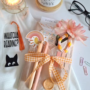 May include: A gift set wrapped in brown paper and tied with a yellow and white checked ribbon. The set includes pens, stickers, and a bracelet. A mug with the words "wife mom boss" is visible, along with a candle and reading-themed accessories.
