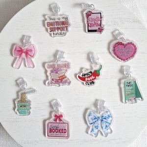 Peut inclure: Une collection de breloques en acrylique transparent avec divers motifs et textes, dont un nœud rose, une pile de livres et les mots "This is my EMOTIONAL SUPPORT kindle". D'autres breloques présentent un piment et un cœur.