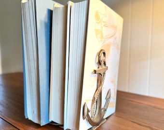 May include: A set of books held together by a nautical-themed bookend. The bookend is a bronze-colored anchor. The books have light blue and cream-colored covers. The books are on a wooden surface.