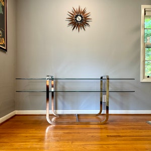 May include: A chrome and glass console table with two shelves. The table has a modern design with a sleek, minimalist aesthetic.