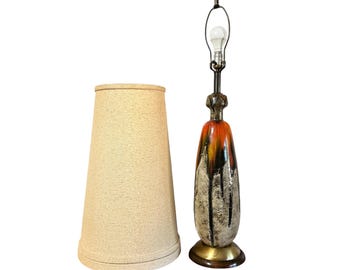 May include: A vintage table lamp with a textured beige lampshade and a tall, ceramic base. The base features a design with orange, black, and white dripped paint, and a brass base. The lamp is turned on with a lightbulb.