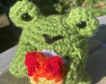 May include: A green crocheted frog holding a rainbow crocheted heart. The frog has black eyes and a black stitched smile.