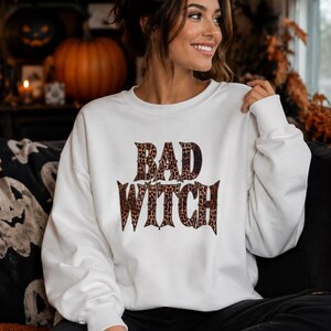 Peut inclure: Un sweat-shirt blanc à manches longues et col rond avec l'inscription "BAD WITCH" imprimée en motif léopard marron et noir sur la poitrine. Le vêtement présente un col, des poignets et un ourlet côtelés, offrant une coupe standard décontractée.