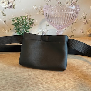 Könnte beinhalten: Eine schwarze Bauchtasche mit einem rechteckigen Beutel und einem elastischen Bund. Der Beutel besteht aus einem glatten Material und ist an einem breiten, schwarzen elastischen Riemen befestigt. Die Bauchtasche ist für das Tragen um die Taille konzipiert.