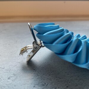May include: A light blue ribbon clip with a silver metal clasp. The ribbon is arranged in a layered, ruffled design. The clip is designed to hold the ribbon in place. The background is a neutral gray.