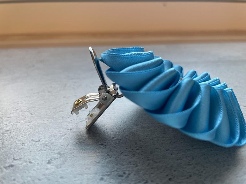 May include: A light blue ribbon clip with a silver metal clasp. The ribbon is arranged in a layered, ruffled design. The clip is designed to hold the ribbon in place. The background is a neutral gray.