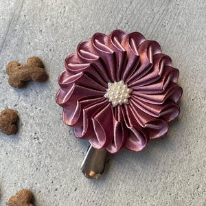 May include: A mauve satin flower hair clip with a pearl center. The flower is made of layered petals and has a silver clip. Several bone-shaped dog treats are scattered around the flower on a gray surface.