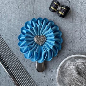 May include: A light blue satin flower-shaped pet grooming accessory with a silver heart-shaped jewel in the center. A black bow with gold polka dots and a silver comb are also visible.