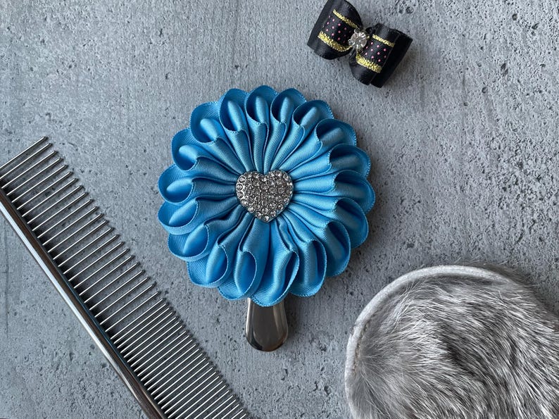 May include: A light blue satin flower-shaped pet grooming accessory with a silver heart-shaped jewel in the center. A black bow with gold polka dots and a silver comb are also visible.
