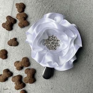 May include: A white satin flower hair clip with a silver rhinestone centre. Scattered around the clip are several brown dog bone-shaped treats. The clip has a black barrette backing.