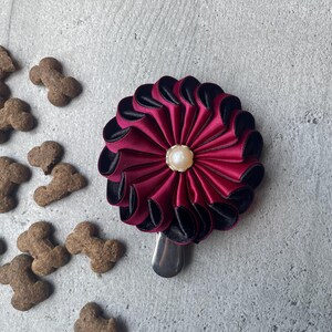 May include: A circular hair accessory featuring layered burgundy and black satin ribbon petals arranged in a flower pattern with a central cream-colored faux pearl bead and a silver-toned metal alligator clip attached to the bottom edge.