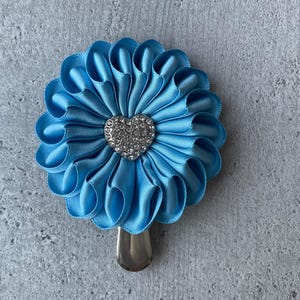 May include: A light blue satin flower hair clip with a silver-colored metal clip. The flower has a heart-shaped rhinestone center. The petals are folded in a ruffled design.