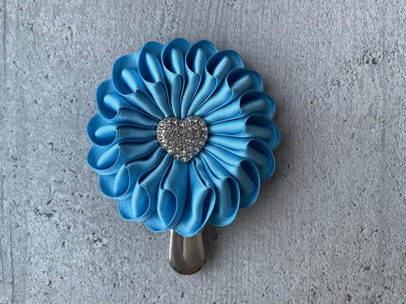 May include: A light blue satin flower hair clip with a silver-colored metal clip. The flower has a heart-shaped rhinestone center. The petals are folded in a ruffled design.