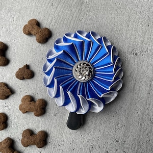 May include: A blue and white rosette-style hair clip with a silver center, next to several brown dog bone-shaped treats. The clip has a black base and is set against a gray textured background.