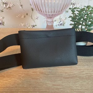 May include: A black rectangular waist bag featuring a smooth, textured surface and a wide, black elastic strap. The bag has a simple, clean design with a horizontal stitched seam across the upper portion, providing a functional accessory for carrying small items.