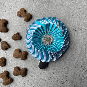 May include: A turquoise and white rosette with a sparkling rhinestone center, next to several bone-shaped dog treats. The rosette is made of pleated fabric and has a black clip on the back. The treats are brown.