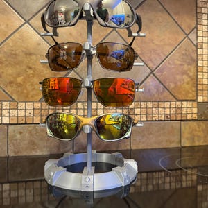 May include: A gray sunglasses display stand holding four pairs of sunglasses with reflective lenses. The stand has a circular base and a vertical support with multiple arms to hold the glasses. The lenses reflect the surrounding environment.