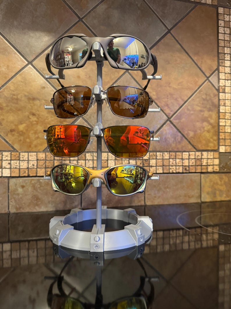 May include: A gray sunglasses display stand holding four pairs of sunglasses with reflective lenses. The stand has a circular base and a vertical support with multiple arms to hold the glasses. The lenses reflect the surrounding environment.