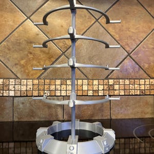 May include: A silver-colored, multi-tiered display stand. The stand has a circular base with a series of vertical supports leading to four curved, semi-circular shelves. Each shelf has small, horizontal extensions. The stand is designed to hold and display items.
