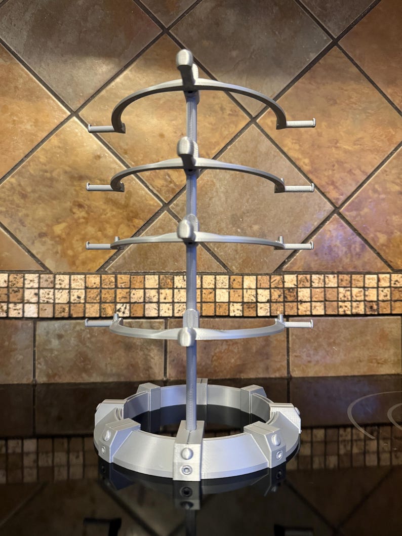 May include: A silver-colored, multi-tiered display stand. The stand has a circular base with a series of vertical supports leading to four curved, semi-circular shelves. Each shelf has small, horizontal extensions. The stand is designed to hold and display items.