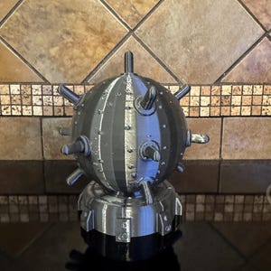 May include: A 3D-printed, gray and black spherical object with protruding spikes and a cylindrical base. The object has a metallic appearance and is likely a decorative item or model.
