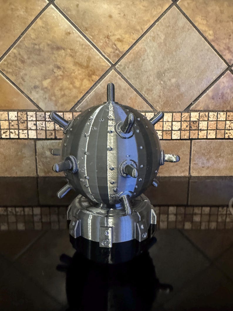 May include: A 3D-printed, gray and black spherical object with protruding spikes and a cylindrical base. The object has a metallic appearance and is likely a decorative item or model.