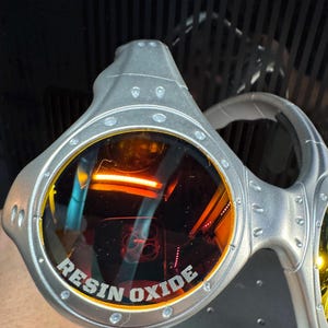 K&ouml;nnte beinhalten: Silberfarbene Brille mit orange get&ouml;nten Gl&auml;sern. Der Text "RESIN OXIDE" ist auf dem Glas aufgedruckt. Die Brille hat ein futuristisches Design mit sichtbaren Nieten und einem gebogenen B&uuml;gel.