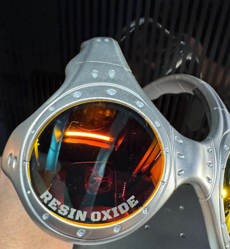 K&ouml;nnte beinhalten: Silberfarbene Brille mit orange get&ouml;nten Gl&auml;sern. Der Text "RESIN OXIDE" ist auf dem Glas aufgedruckt. Die Brille hat ein futuristisches Design mit sichtbaren Nieten und einem gebogenen B&uuml;gel.