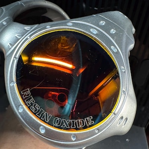 K&ouml;nnte beinhalten: Silberfarbene Sonnenbrille mit orange get&ouml;nten Gl&auml;sern. Der Text "RESIN OXIDE" ist auf dem Rahmen aufgedruckt. Die Sonnenbrille hat ein einzigartiges, eckiges Design.