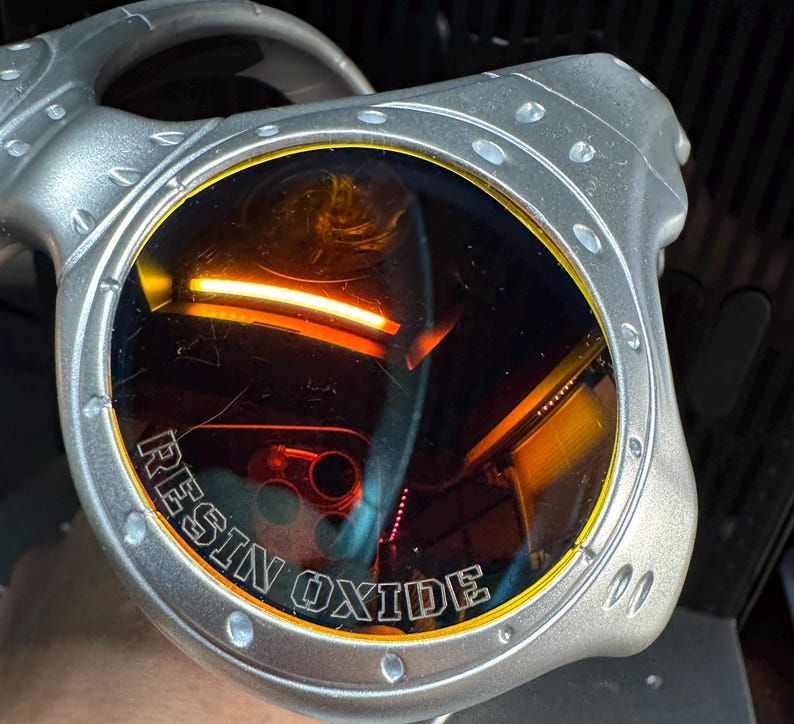 K&ouml;nnte beinhalten: Silberfarbene Sonnenbrille mit orange get&ouml;nten Gl&auml;sern. Der Text "RESIN OXIDE" ist auf dem Rahmen aufgedruckt. Die Sonnenbrille hat ein einzigartiges, eckiges Design.