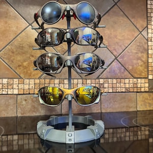 May include: A display stand holding four pairs of sunglasses. The sunglasses have various frame colors, including silver, black, and gold, with reflective lenses. The stand is gray and has a circular base.