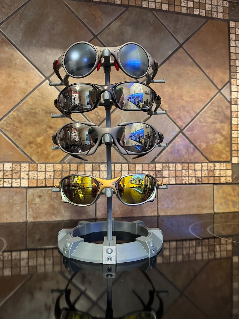 May include: A display stand holding four pairs of sunglasses. The sunglasses have various frame colors, including silver, black, and gold, with reflective lenses. The stand is gray and has a circular base.