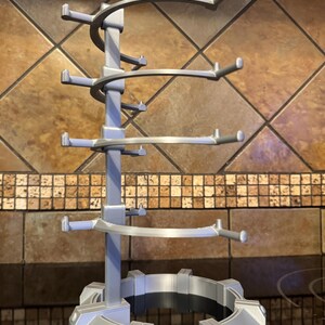 May include: A silver-colored, multi-tiered mug tree with a circular base and multiple arms extending outwards to hold mugs. The tree has a spiral design and is made of a light-colored plastic material. The mug tree is designed to hold multiple mugs.
